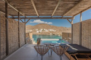 A covered outdoor seating area with a pool, overlooking a mountainous landscape and a coastal town. - Amal Kythnos Suites (Kithnos)