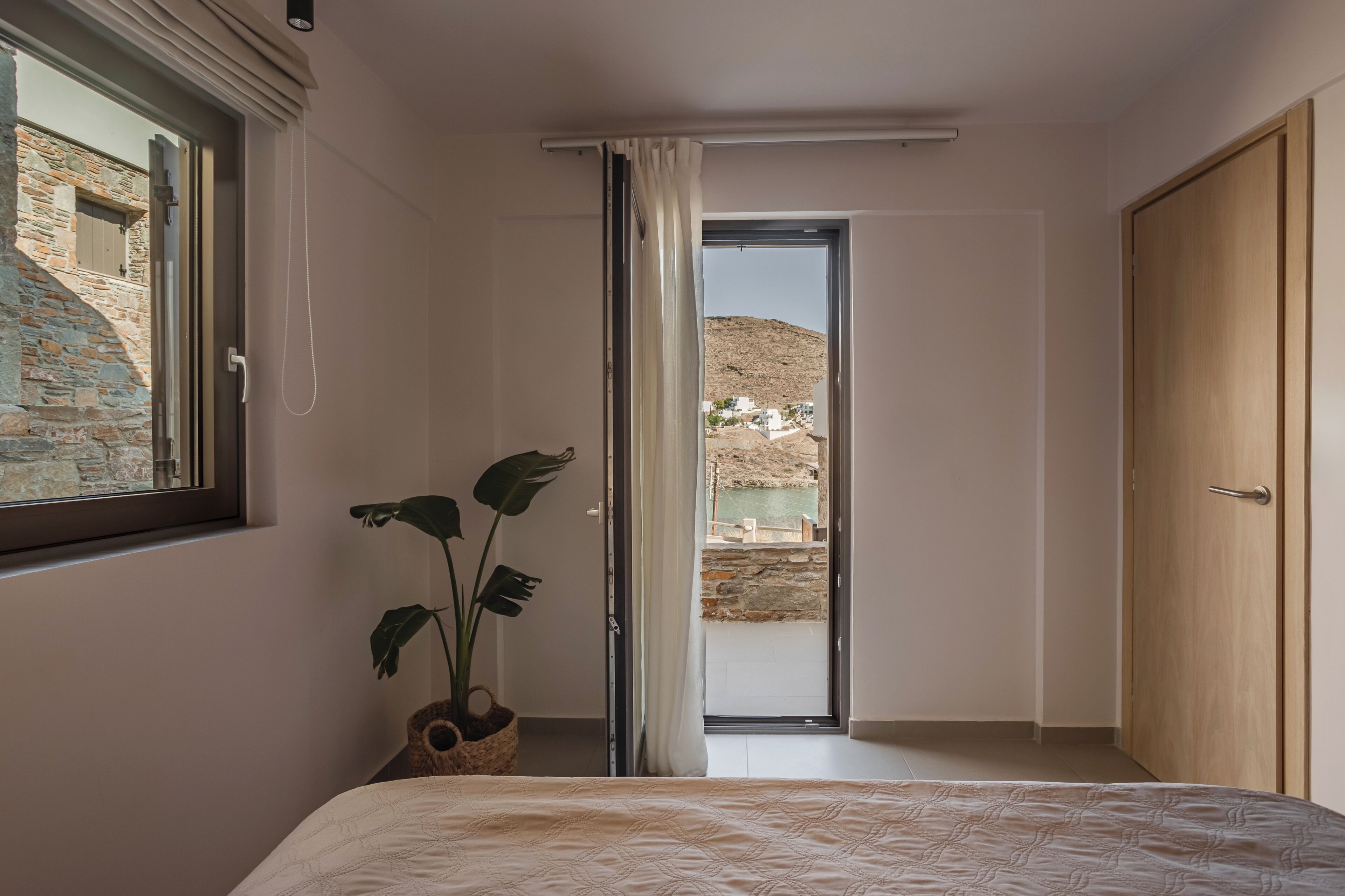 Un dormitorio con cama, planta en maceta, puerta de madera y vista a un paisaje montañoso a través de una puerta abierta.