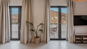 A modern living room with a large TV, a dining area with a table and chairs, and a balcony with a view of a hillside.
