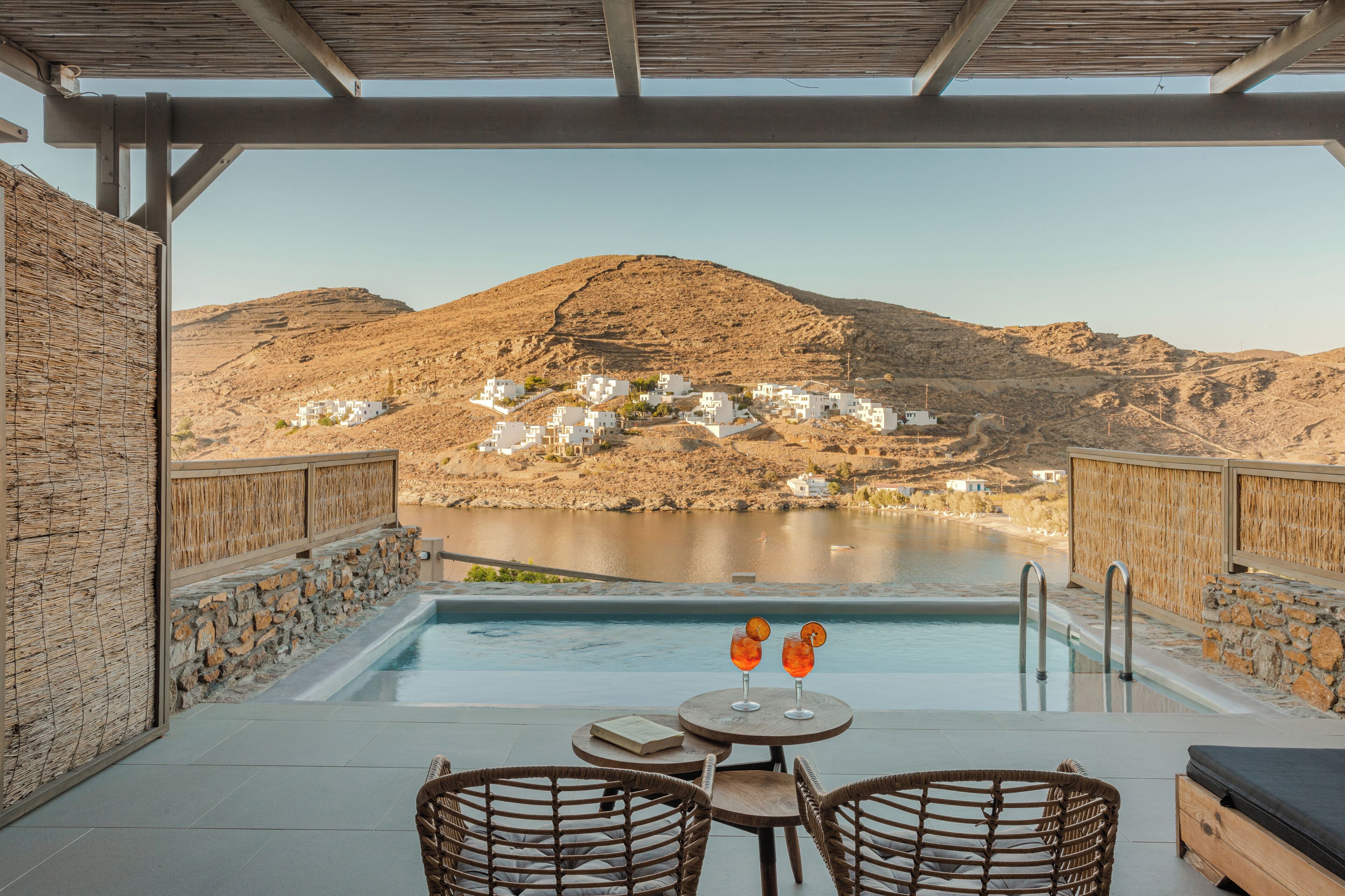 Un espace extérieur avec une piscine, des fauteuils en osier et une table, offrant une vue sur une colline parsemée de bâtiments blancs et un plan d’eau.