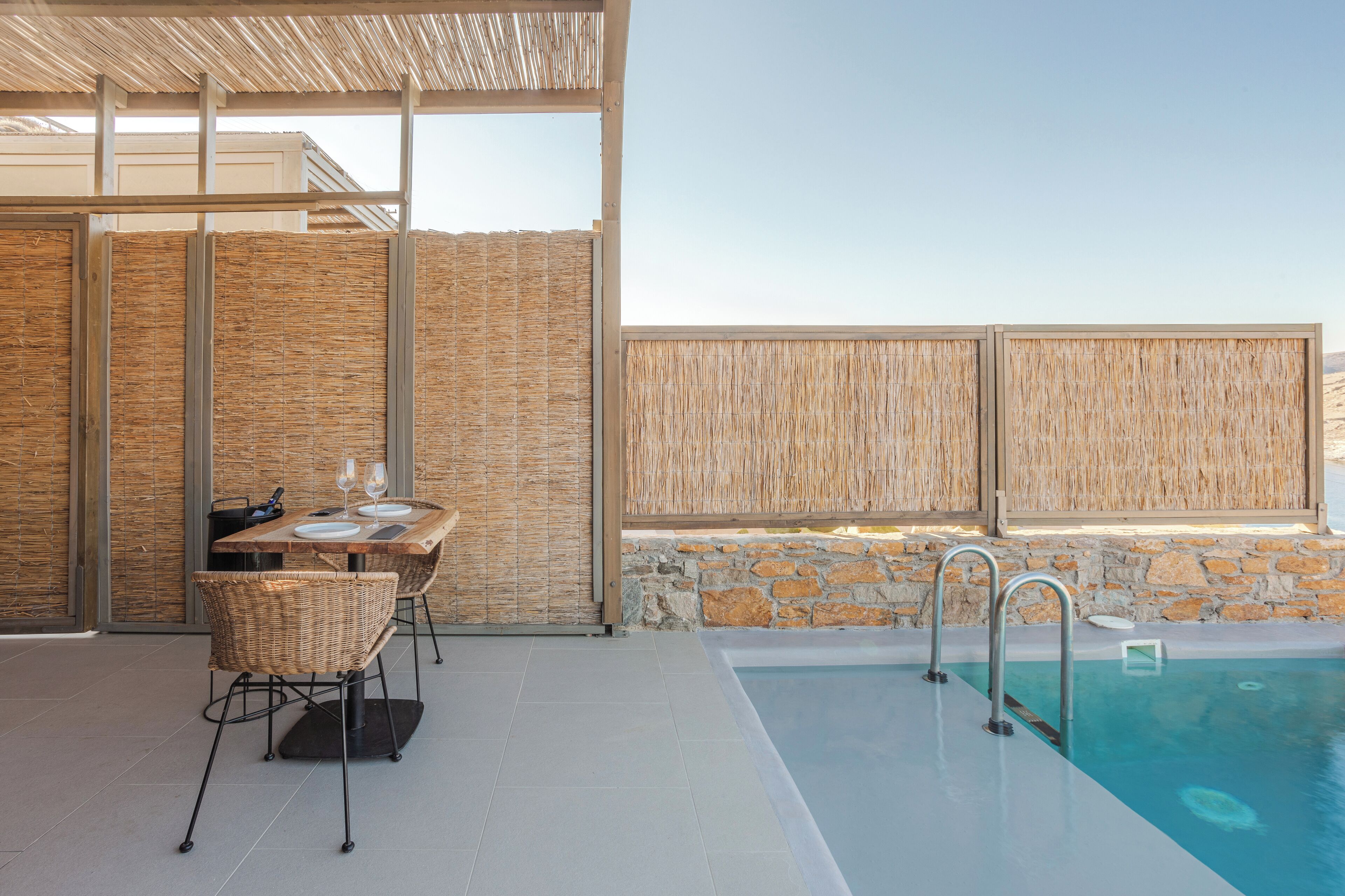 A poolside area with a table and chairs, a stone wall, and a pool with a ladder.