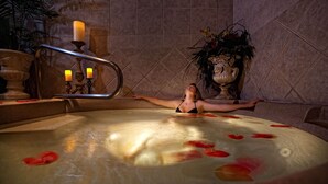 Eine Frau entspannt sich in einer Hot-Tub, während Rosenblätter im Wasser treiben, umgeben von einer gefliesten Wand und einer Zierpflanze.