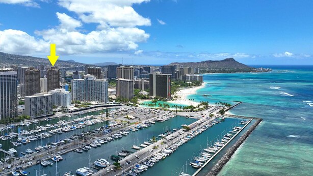 A marina with numerous boats docked, a beachfront, and high-rise buildings in the background.