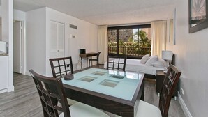 A hotel room with a glass-top dining table, a bed with white linens, a desk with a chair, and a window with a view of palm trees.