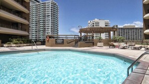 A swimming pool with a lifeguard stand, surrounded by high-rise buildings and a covered area with lounge chairs.