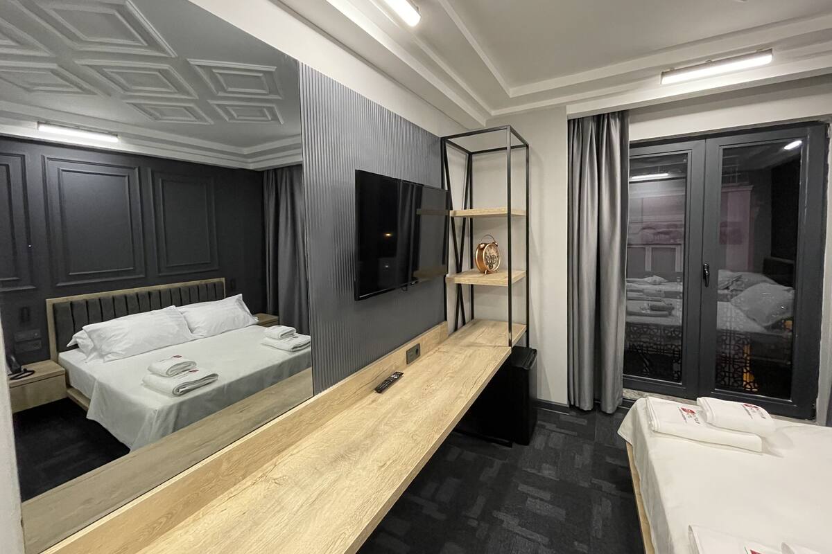A modern hotel room with a large bed, a bedside table, a TV mounted on the wall, and a large mirror.