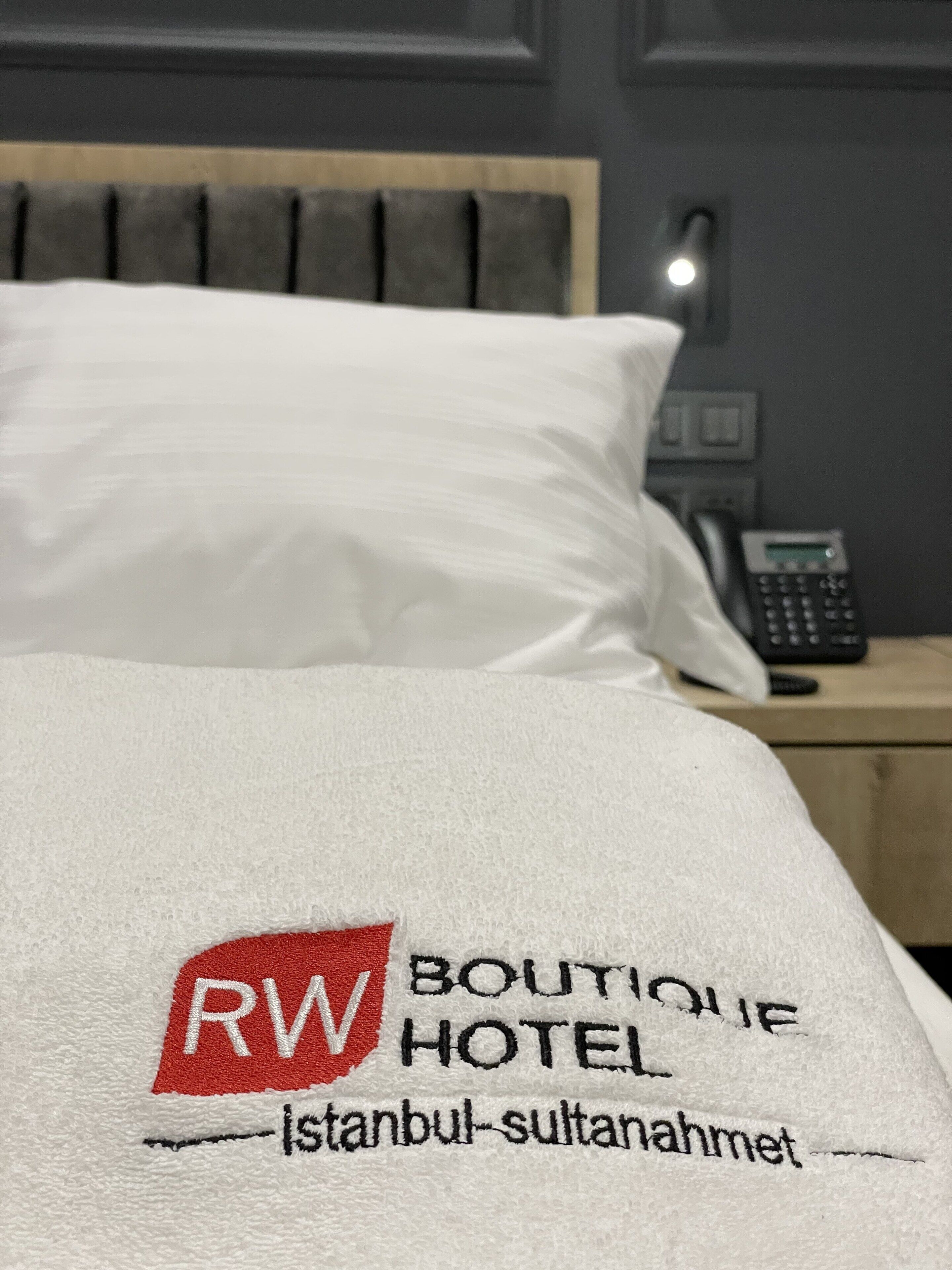 Una cama con almohada blanca y manta que muestra el logo y el nombre del RW Boutique Hotel en Estambul-Sultanahmet.