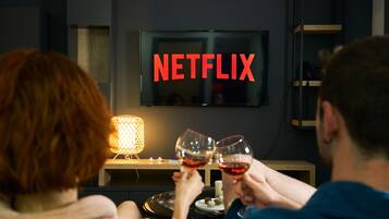 Una habitación con una televisión mostrando el logotipo de Netflix, una mesa con una vela y dos personas sosteniendo copas de vino.