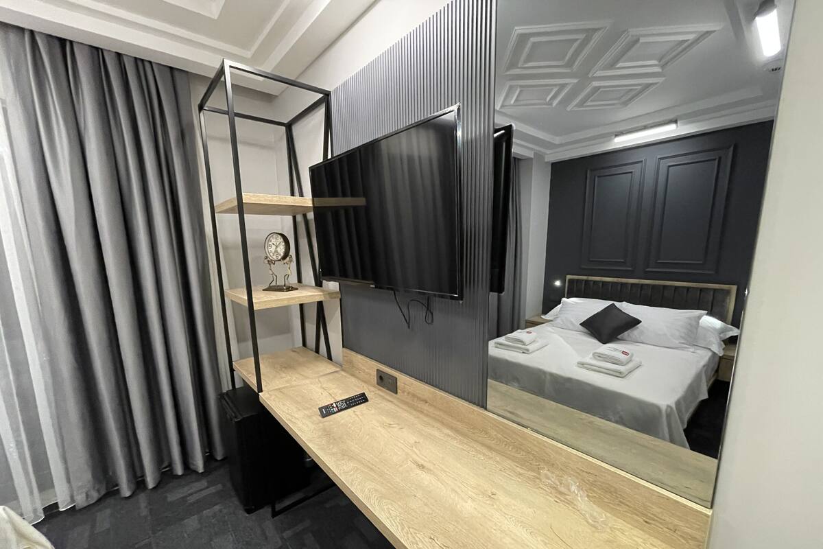 A modern hotel room with a wooden desk, a flat-screen TV mounted on the wall, a bed with white linens, and a large mirror reflecting the room.
