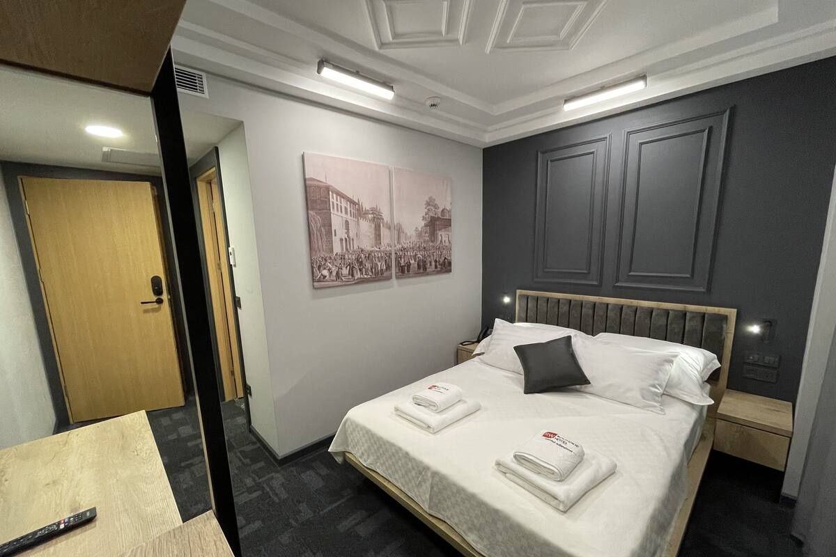 A hotel room with a bed, a nightstand, a wall-mounted lamp, and a door.