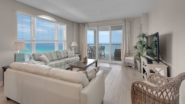A spacious living room with a large sofa, a coffee table, and a television. There is a balcony with a view of the ocean. - Beach Colony East 15A- 3BD/3BA Gulf front w/Beach Service & Onsite Management (Navarre Beach)