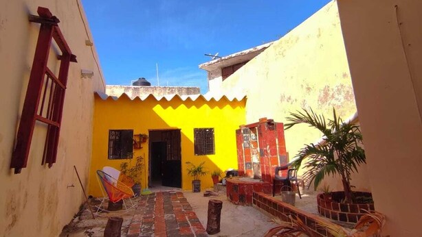 A narrow alleyway with a yellow building featuring a black door and windows. There are potted plants and a small seating area with chairs. - LET'S ROLA (Campeche)