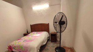 A small room with a bed, a nightstand, a bedside table, a standing fan, and a green chair. - LET'S ROLA (Campeche)