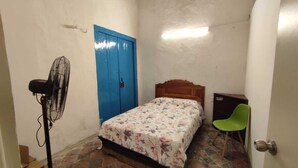 A small, narrow room with a bed, a blue door, a fan, a green chair, and a tiled floor. - LET'S ROLA (Campeche)