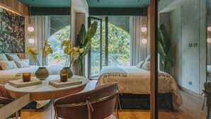 Ein modernes Hotelzimmer mit großem Fenster, einem Bett, einem Sessel, einem Tisch mit Büchern und einer Kerze sowie einer Zierpflanze.