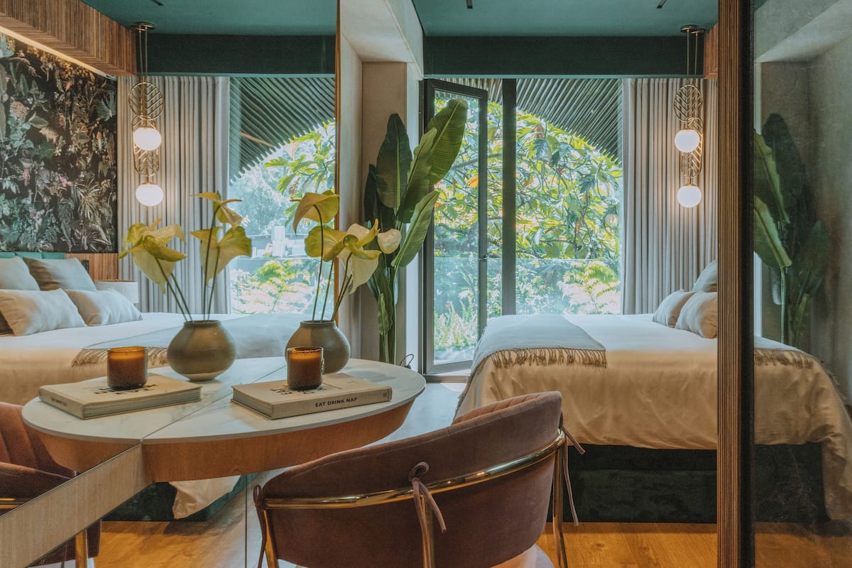 Une chambre d’hôtel moderne avec une grande fenêtre, un lit, une chaise, une table avec des livres et une bougie, ainsi qu’une plante décorative.