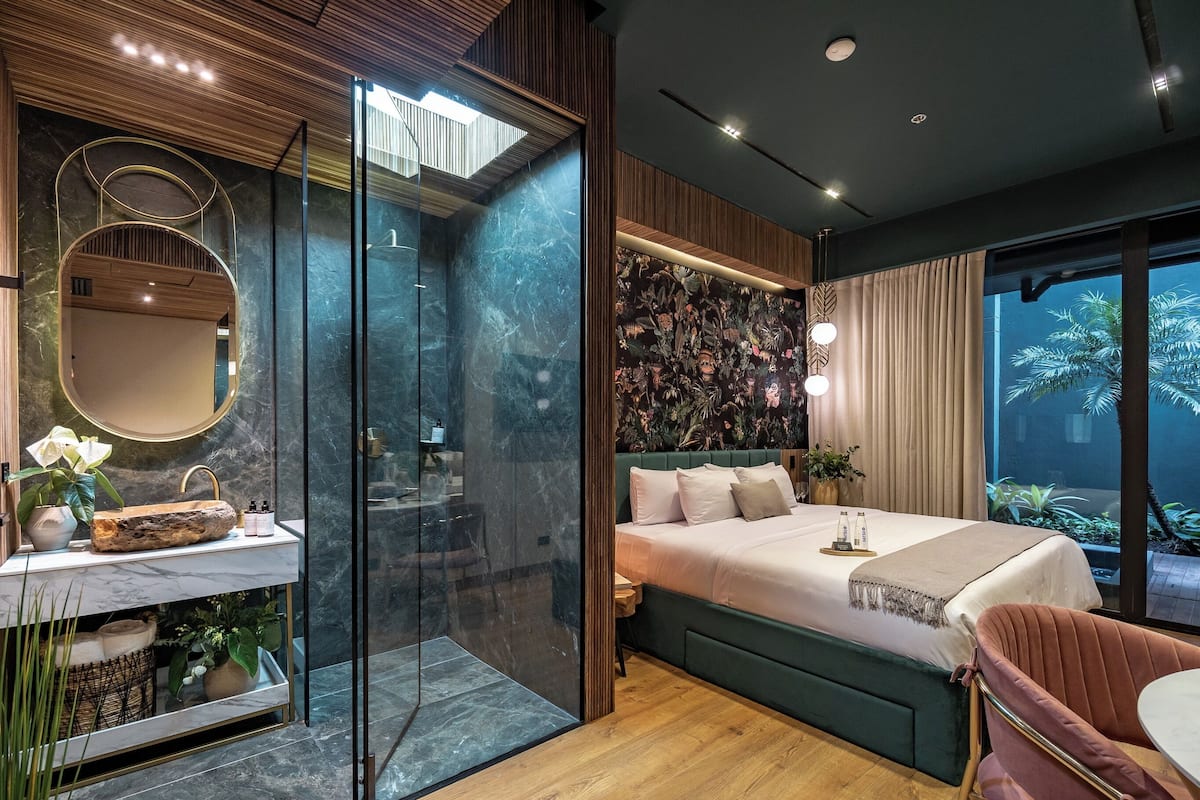 Une chambre moderne dotée d’une douche en verre, d’un grand lit avec une tête de lit verte et d’une vue sur un jardin tropical.