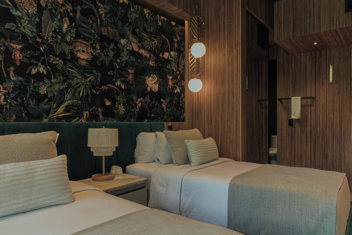 Une chambre d’hôtel avec deux lits, une tête de lit verte et un mur orné d’une fresque représentant des plantes tropicales, ainsi que deux lampes fixées au mur.