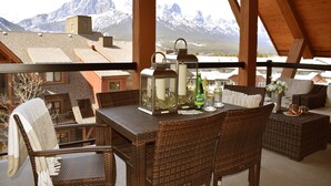 A balcony with wicker furniture, a table with a bottle, and a view of snow-capped mountains. - Extraordinary Mountain Penthouse, Spectacular Views, Pool & Hot Tub, Park Pass (Canmore)