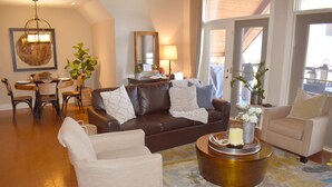 A spacious living room with a brown leather sofa, beige armchairs, and a coffee table. There is a dining area with a wooden table and chairs, and a large window providing natural light. - Extraordinary Mountain Penthouse, Spectacular Views, Pool & Hot Tub, Park Pass (Canmore)