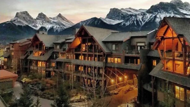 A lodge with multiple wooden buildings, a central courtyard, and a view of snow-capped mountains in the background. - Extraordinary Mountain Penthouse, Spectacular Views, Pool & Hot Tub, Park Pass (Canmore)