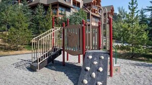 A playground with a wooden slide and climbing structures, surrounded by trees and a building in the background. - Extraordinary Mountain Penthouse, Spectacular Views, Pool & Hot Tub, Park Pass (Canmore)