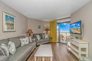 A living room with a sofa, a coffee table, a TV, and a balcony with a view.
