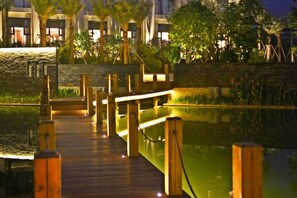 A wooden boardwalk leading to a building with a stone facade, surrounded by greenery and a body of water. - Lotus Villa Hotel (Liuzhou)