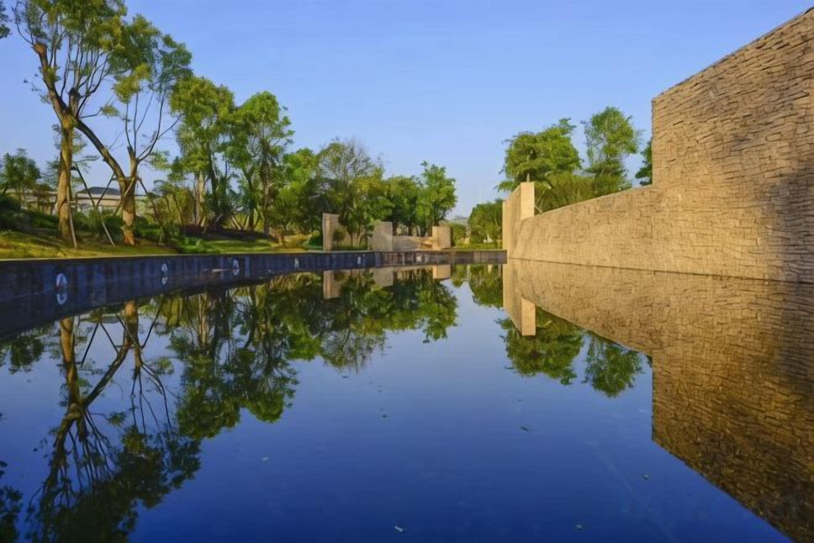 A calm water body reflecting a brick wall and trees.