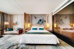 Een hotelkamer met een groot bed, een bank en een bureau. Er hangen schilderijen aan de muur en er is een groot raam.