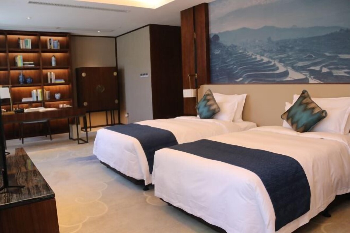 Een hotelkamer met twee bedden, een boekenkast, een bureau en een landschapsschilderij.