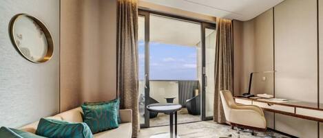 A modern hotel room with a sofa, a small round table, a desk with a chair, and a view through a glass door.