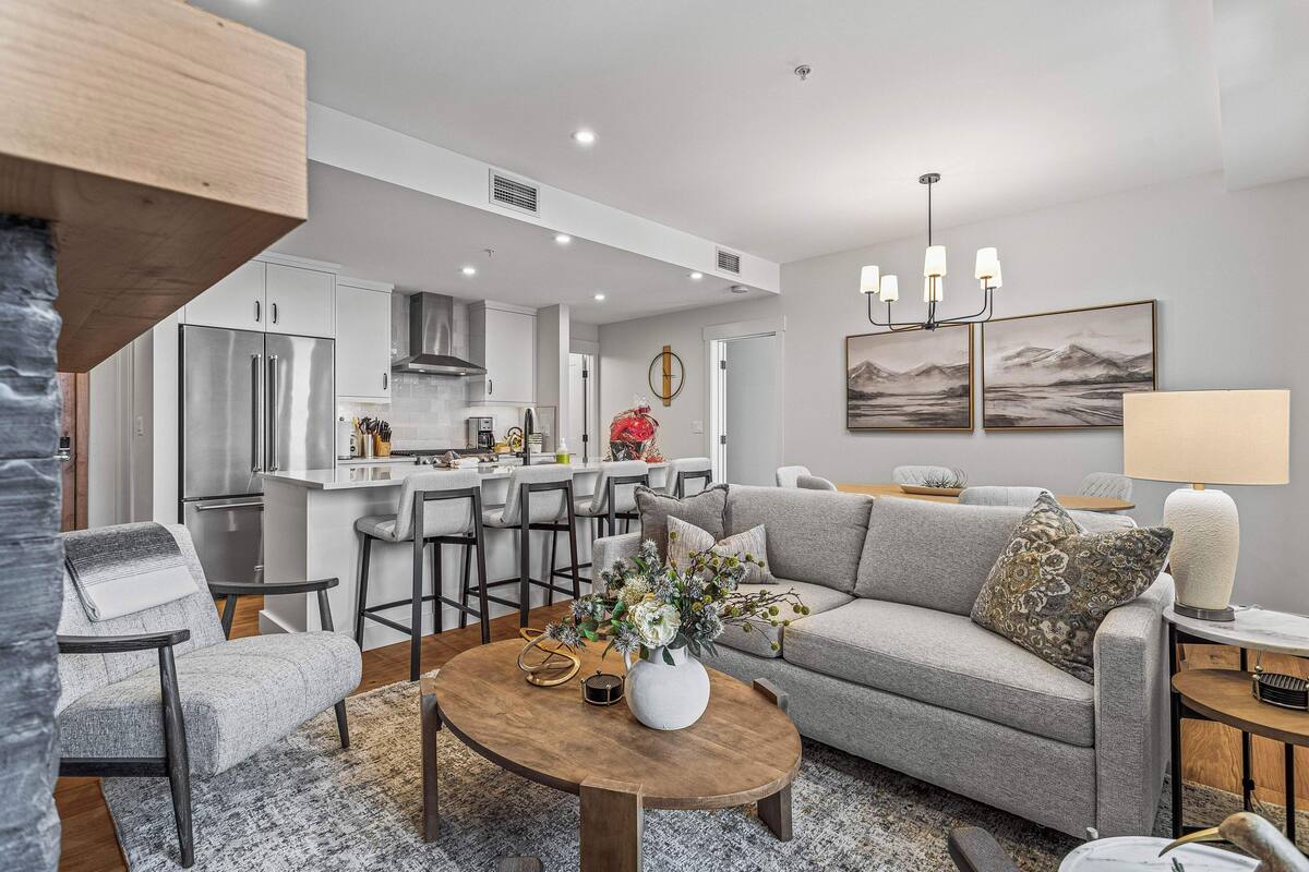 A modern living room with a grey sofa, a wooden coffee table, and a kitchen in the background.