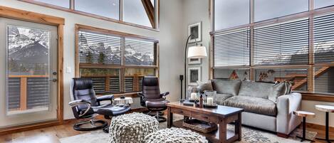 A spacious living room with large windows, a leather chair, a sofa, ottomans, and a coffee table.