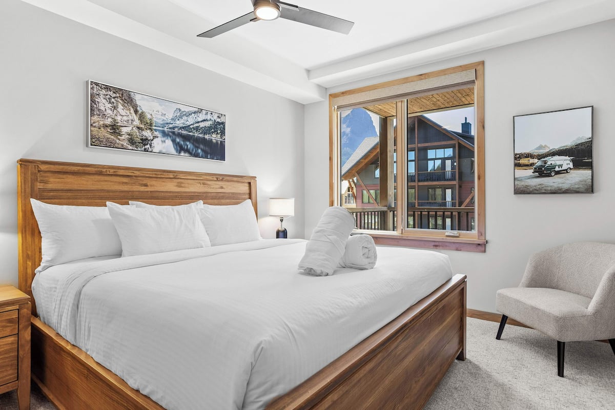 A bedroom with a wooden bed, a window with a view of a mountain cabin, and a chair.