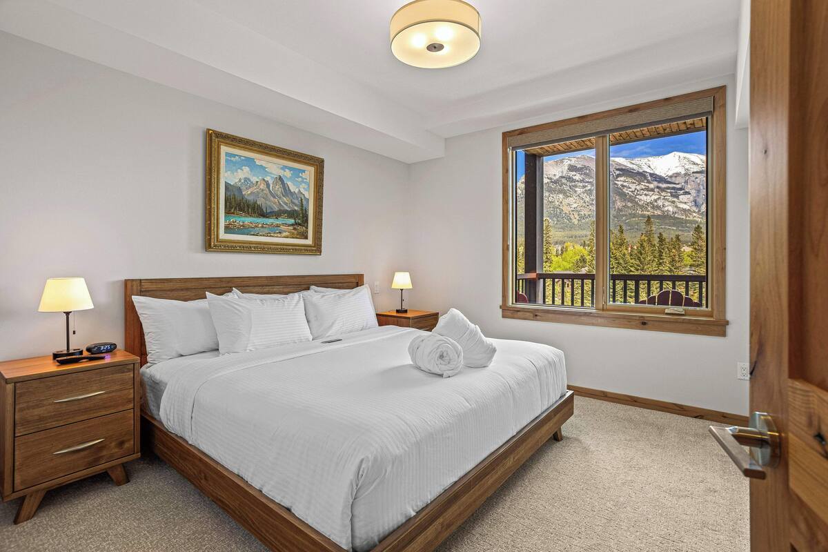 A bedroom with a large bed, wooden headboard, two bedside tables with lamps, a painting on the wall, and a window with a mountain view.