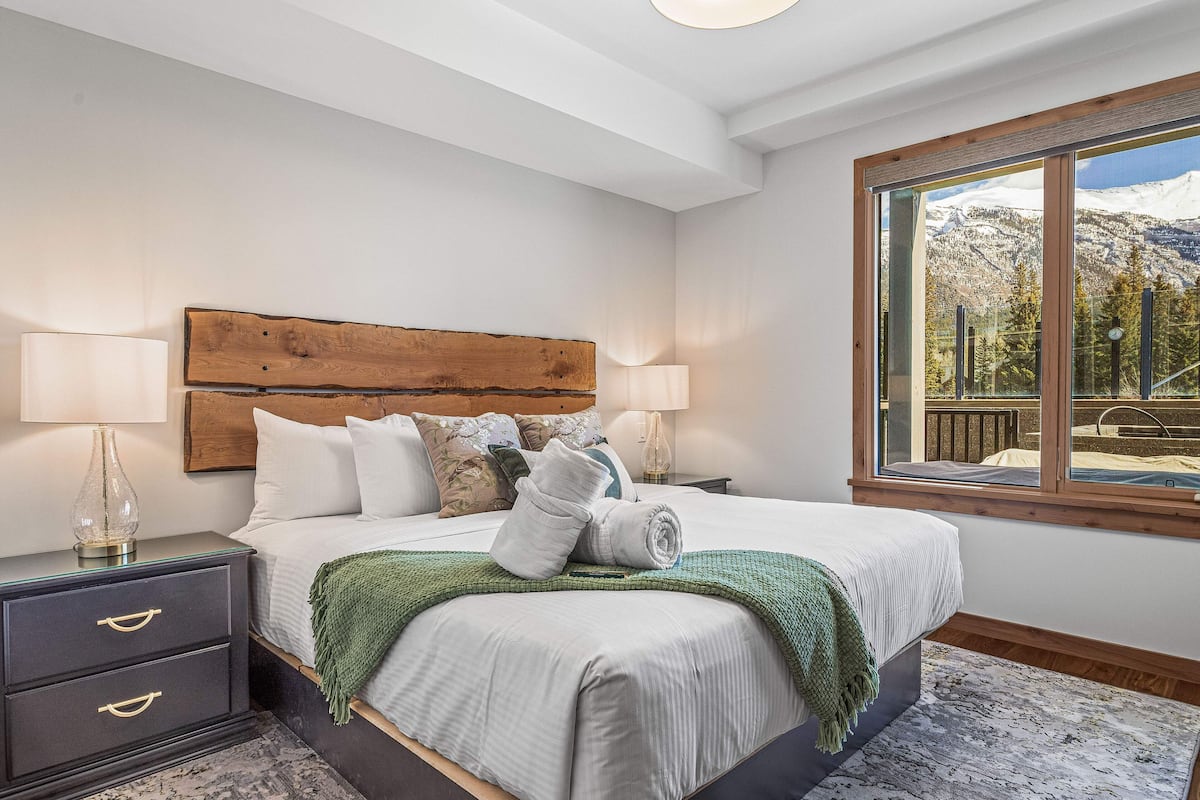 A neatly made bed with white linens, a wooden headboard, and a view of snow-capped mountains through a large window.