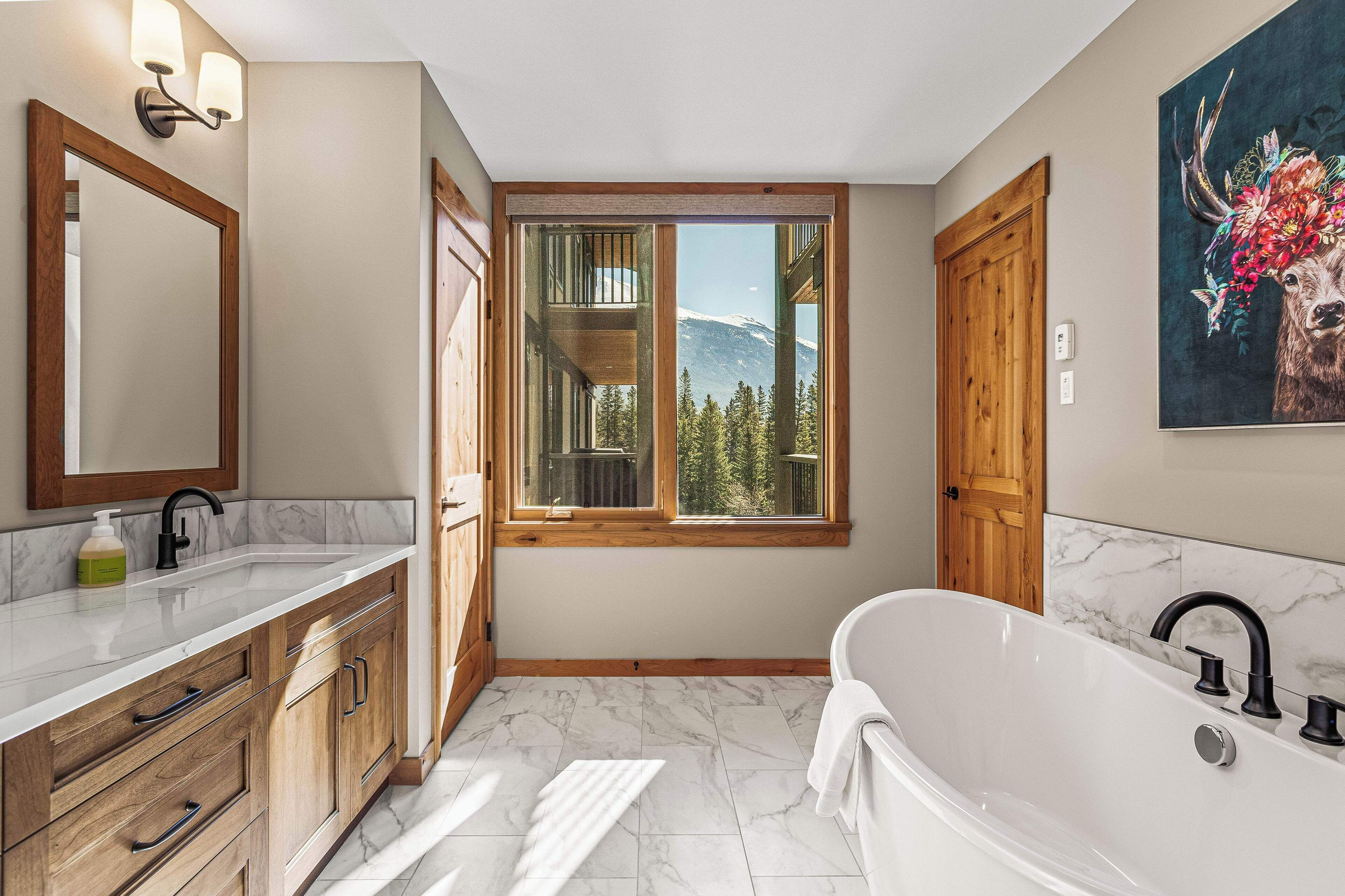 Un baño con un amplio ventanal, armarios de madera, una bañera blanca y un cuadro de un ciervo.