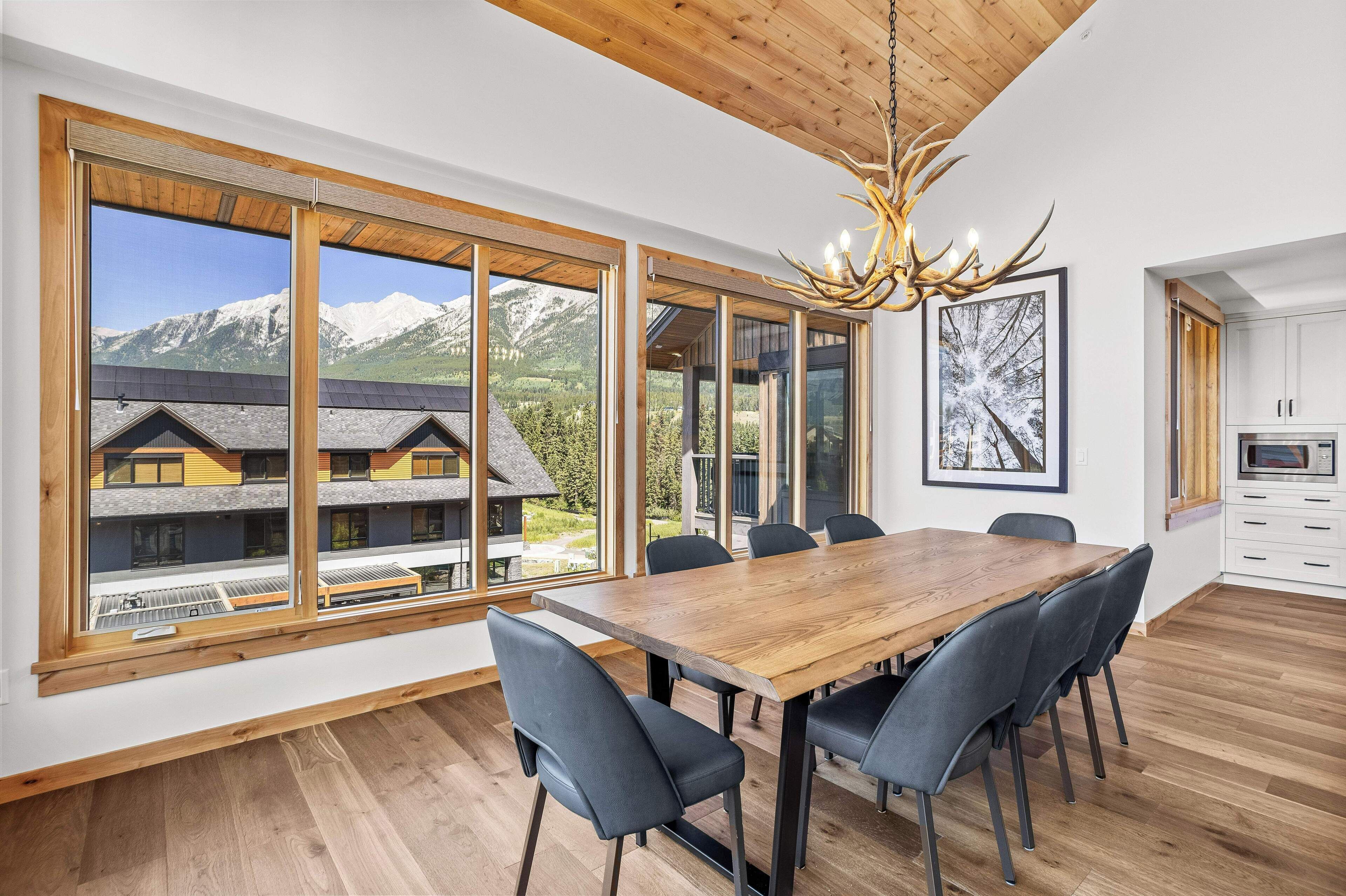 A modern dining area with a large wooden table and chairs, large windows offering mountain views, and a chandelier.
