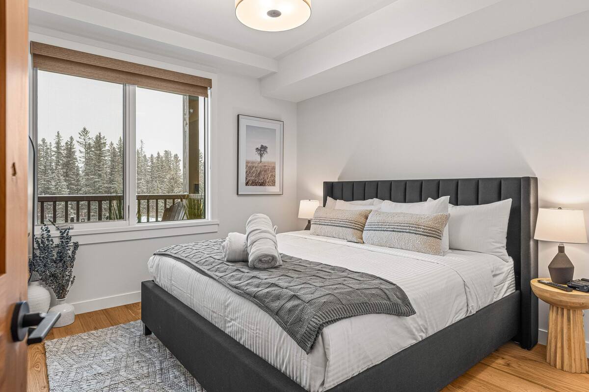 A bedroom with a large bed, a window with a view of trees, a bedside table with a lamp, and a framed picture on the wall.