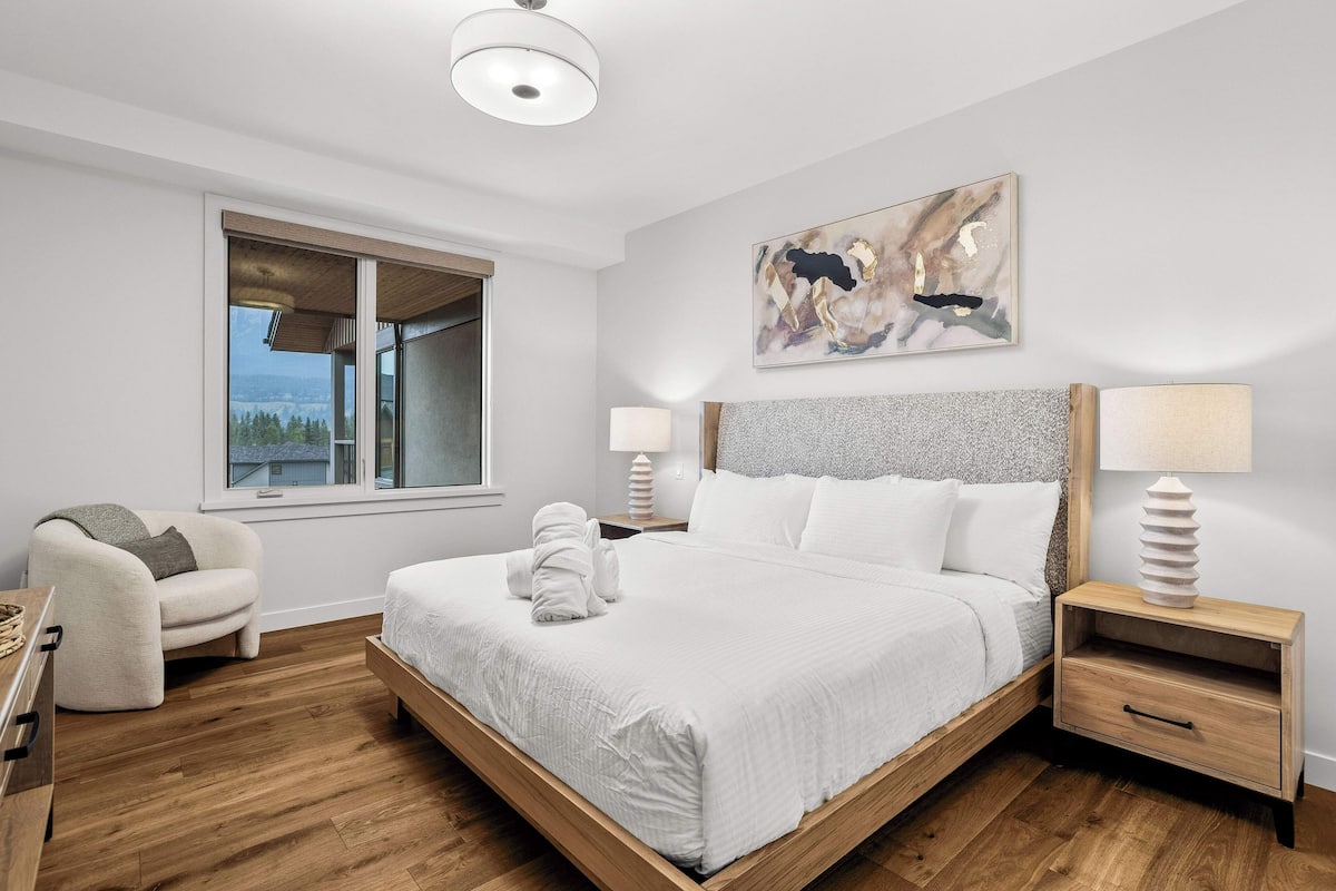 A modern bedroom with a large bed, bedside lamps, a view through a window, and a painting on the wall.