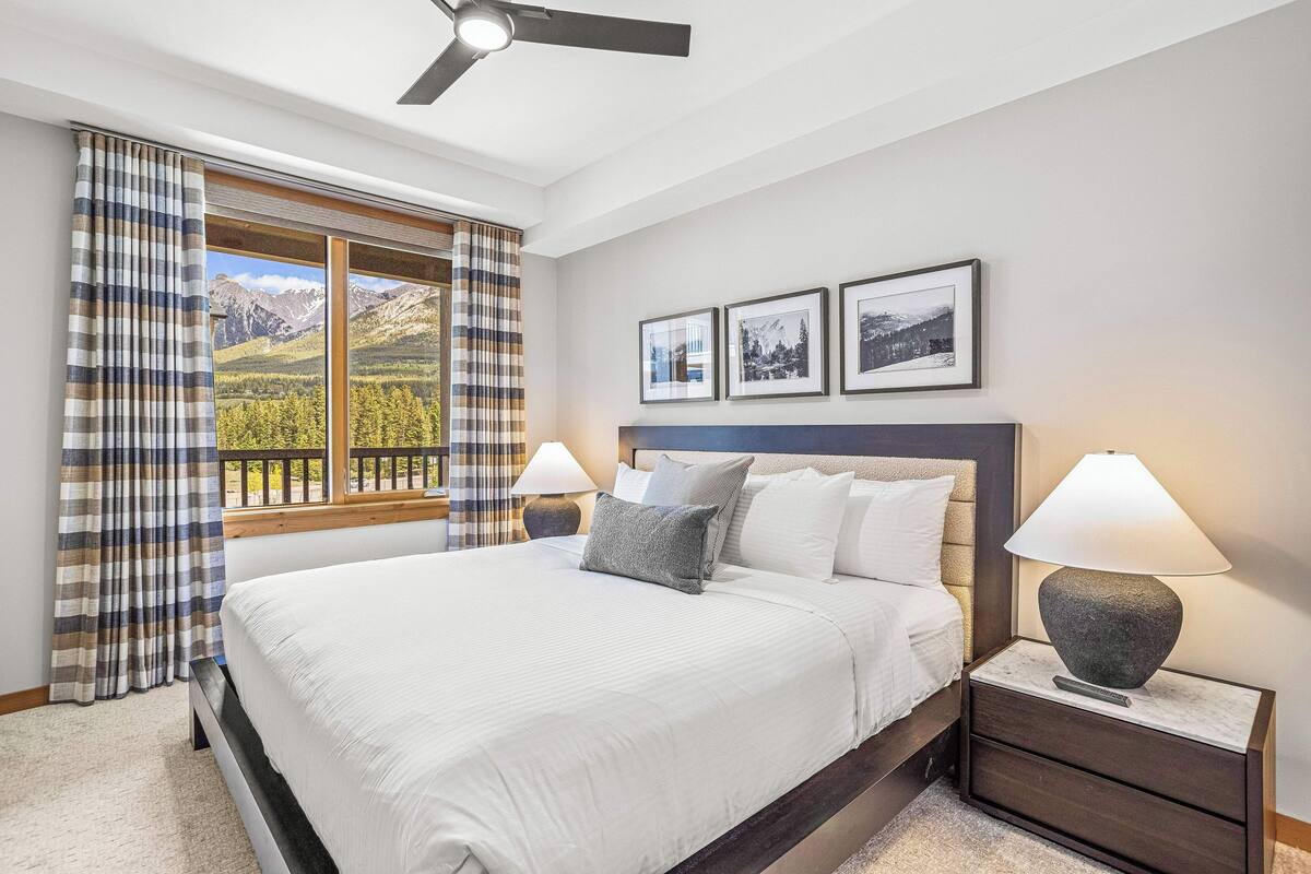 A bedroom with a large bed, two bedside tables with lamps, a ceiling fan, and a window with curtains.