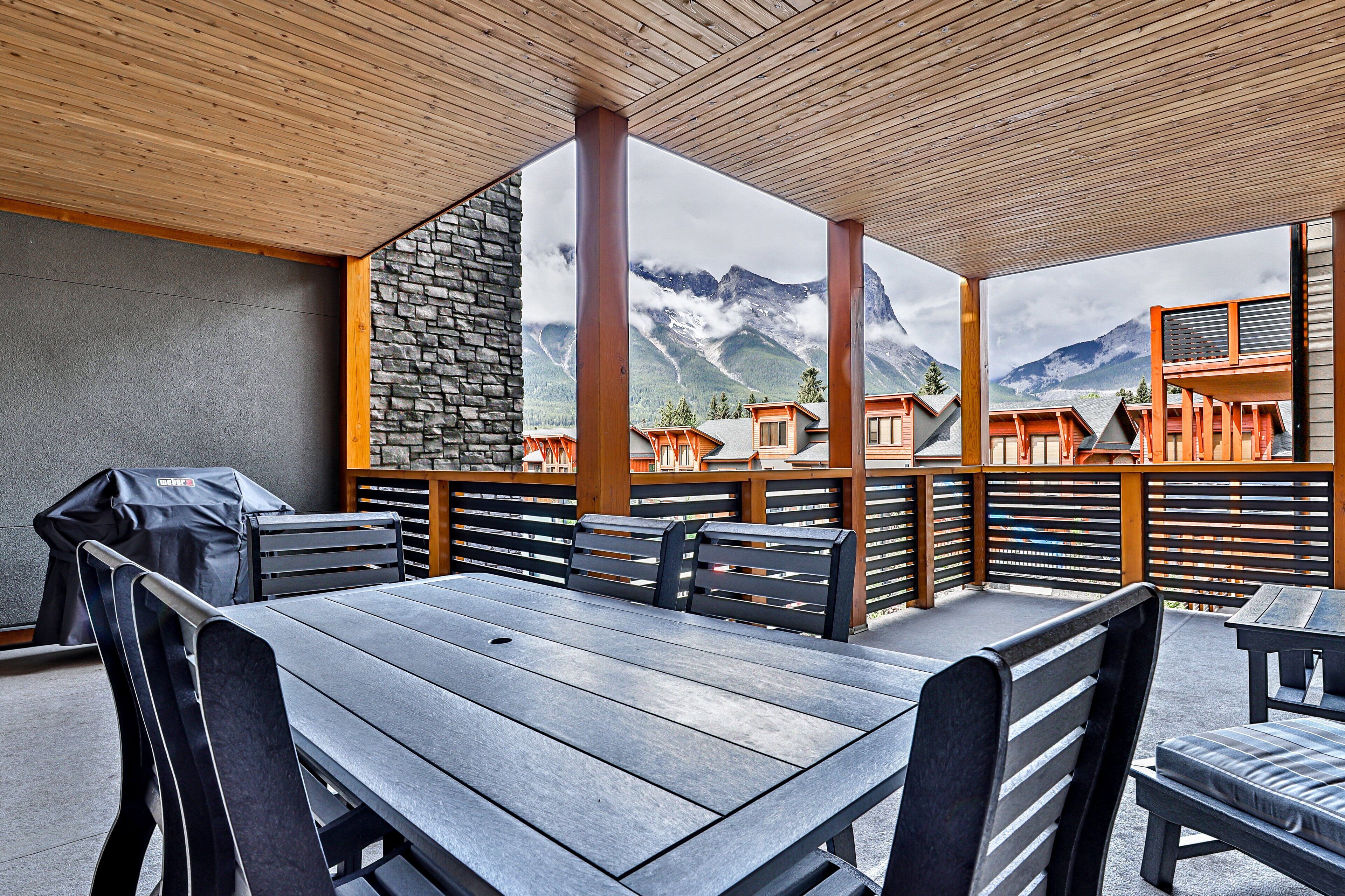 A covered outdoor seating area with a table and chairs, a grill, and a view of mountains and buildings.