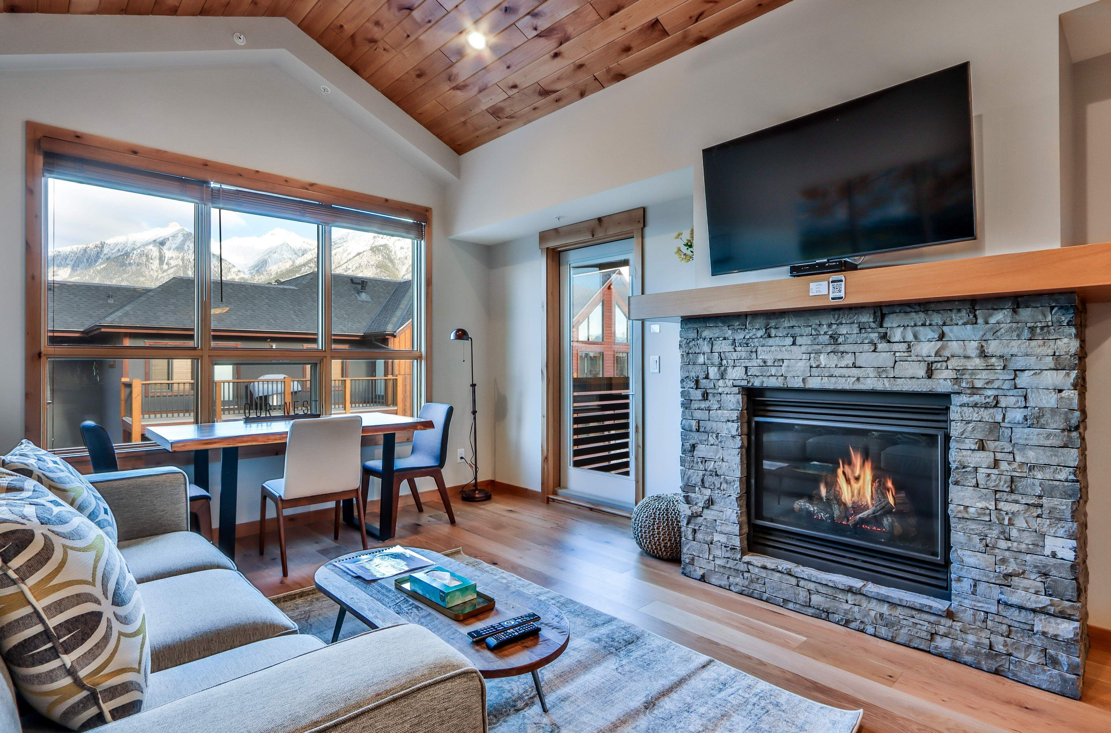 A living room with a stone fireplace, a flat-screen TV, and a dining area with a view of mountains.