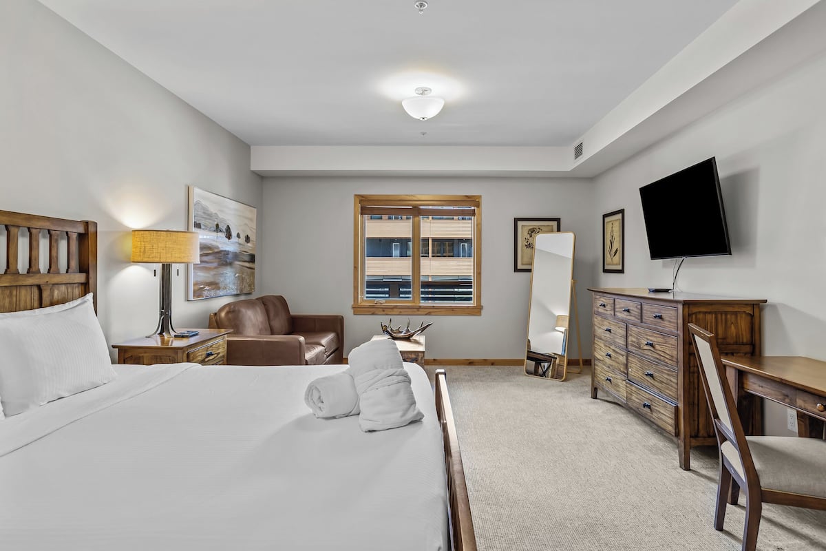 A hotel room with a bed, a desk, a chair, a television, a lamp, a leather chair, a wooden dresser, a window with blinds, and artwork on the walls.