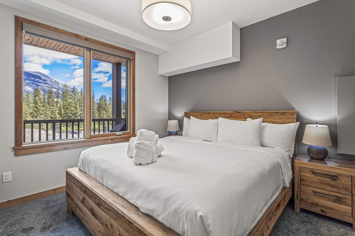 A neatly made bed with a wooden headboard, a nightstand with a lamp, and a window with a view of trees and mountains.