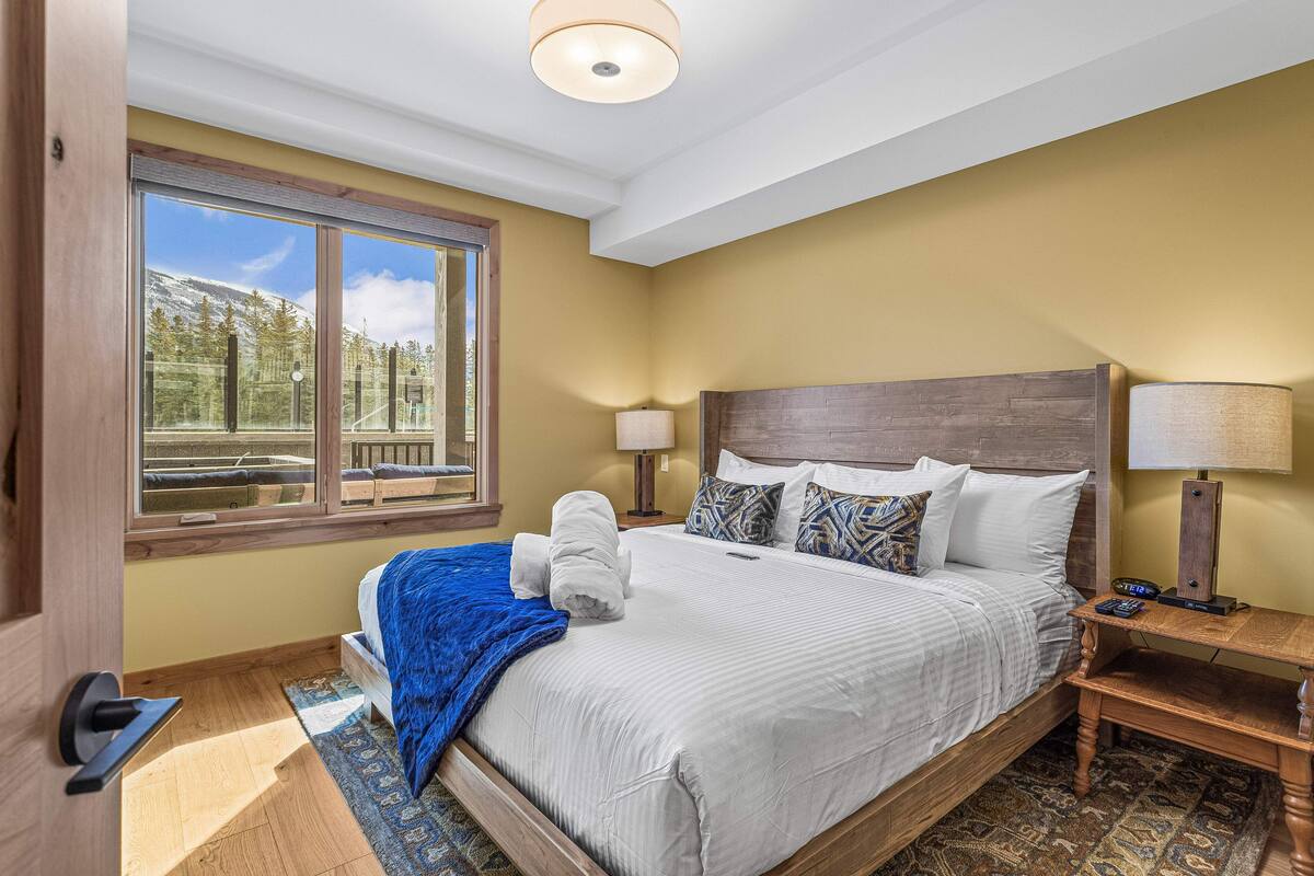 A hotel room with a large bed, a wooden headboard, two bedside lamps, a nightstand, a window with a view of trees and mountains, and a blue throw blanket on the bed.