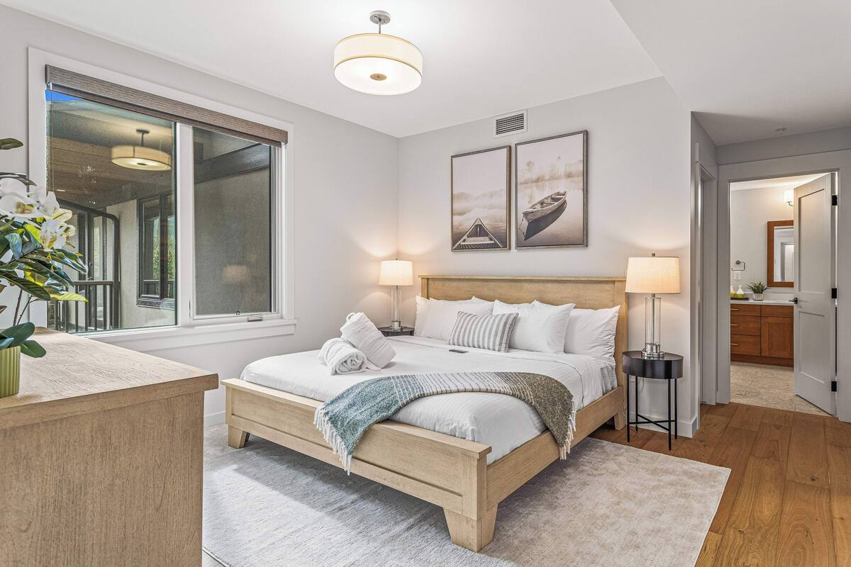 A modern bedroom with a large window, a wooden bed, bedside lamps, and artwork on the wall.