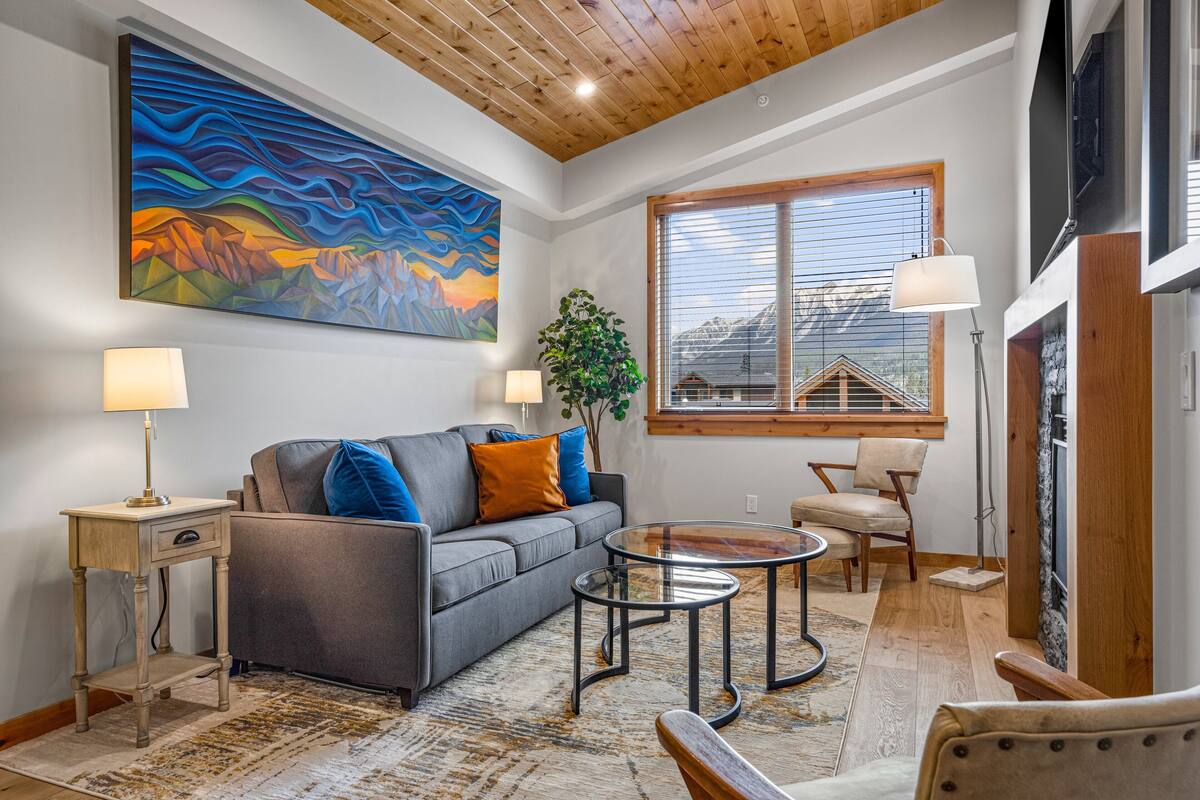 Una sala de estar moderna con un sofá gris, cojines azules y naranjas, una mesa de centro de vidrio y un cuadro abstracto grande en la pared.