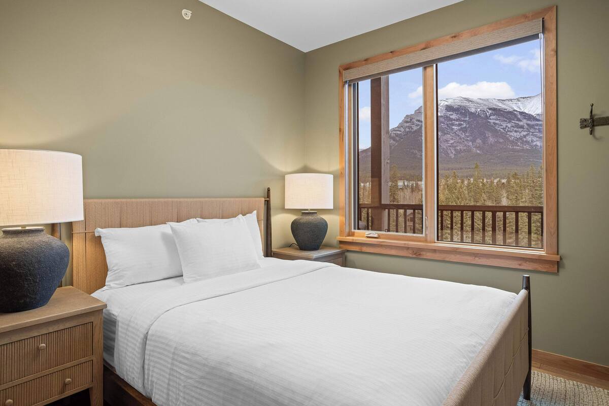A neatly made bed with white linens, a wooden headboard, and a nightstand with a lamp and a vase. A large window with a wooden frame offers a view of a mountain and trees.